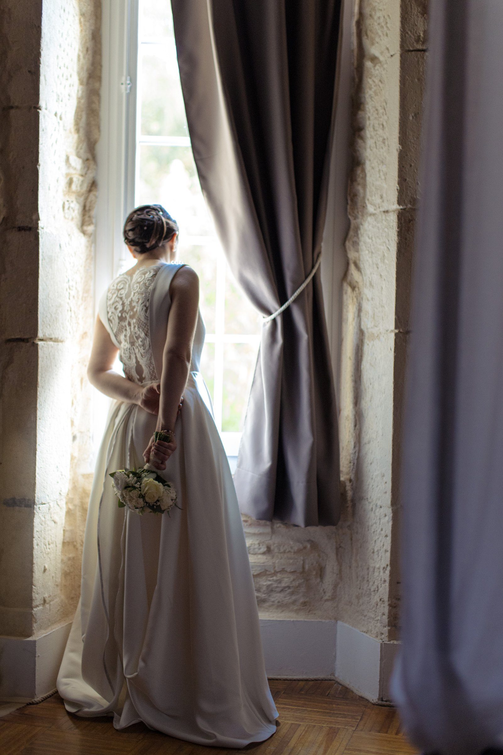 La mariée de dos devant une grande fenêtre, bouquet à la main